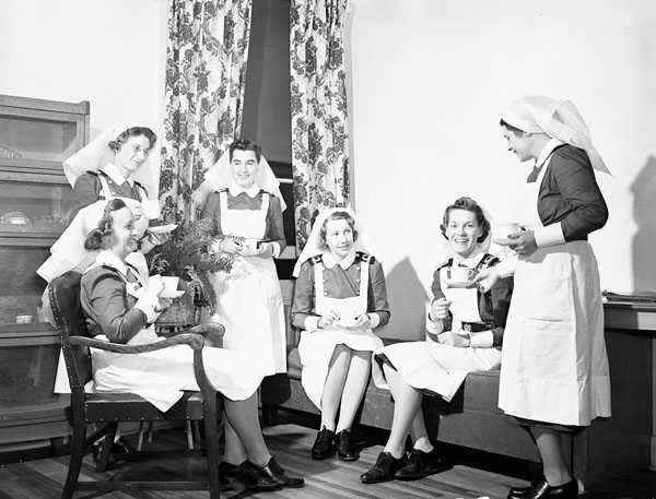 Black and white photograph. 6 women in nursing uniforms (blue dress with white aprons and headdresses) sit/stand in a room. There is a piece of wooden furniture and floral curtains visible. They hold teacups and chat with each other.